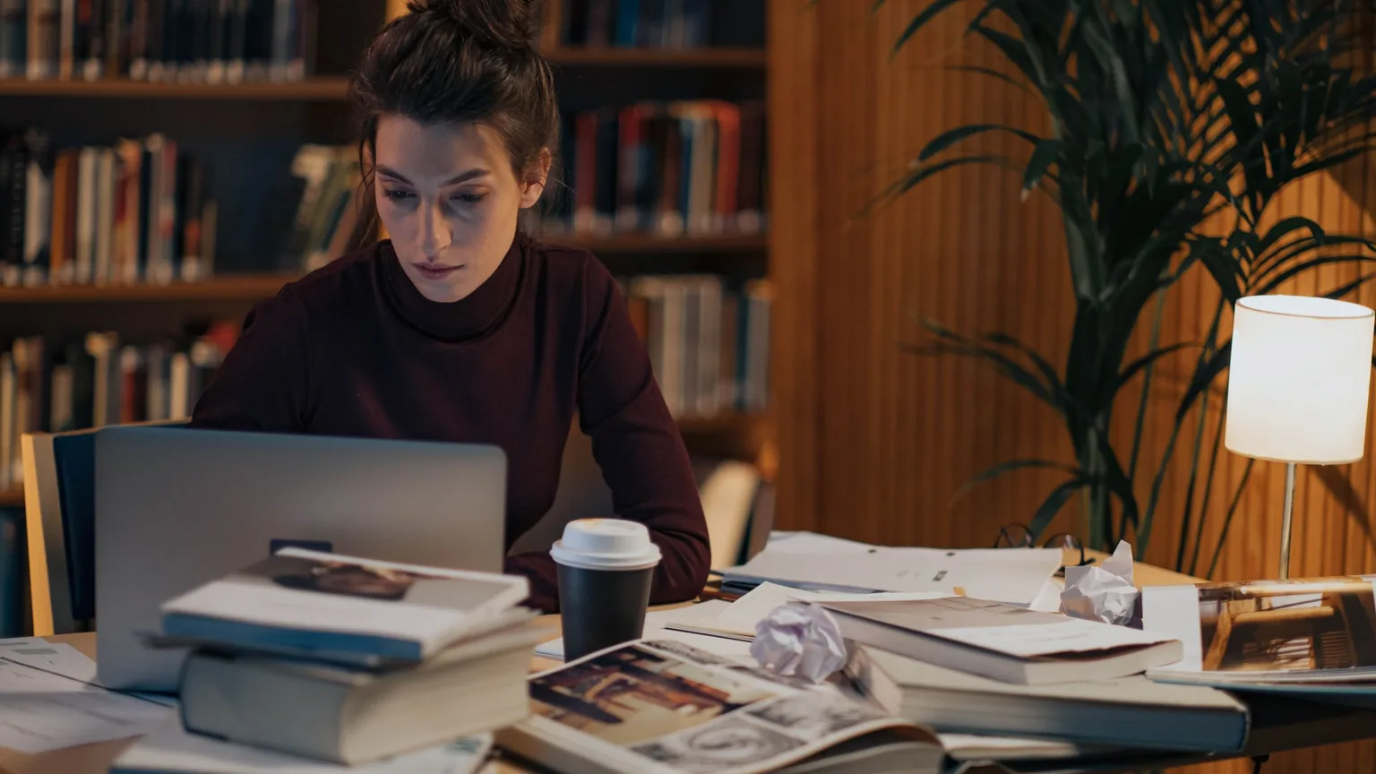 A club secretary working late at a laptop in the library, books and papers spread across the desk