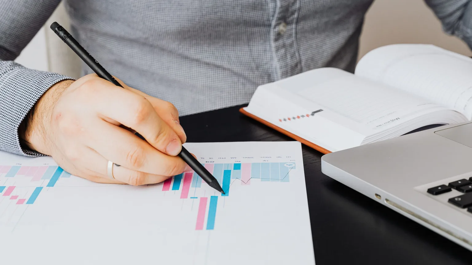 A hand marking up a printed bar chart beside a notebook and laptop