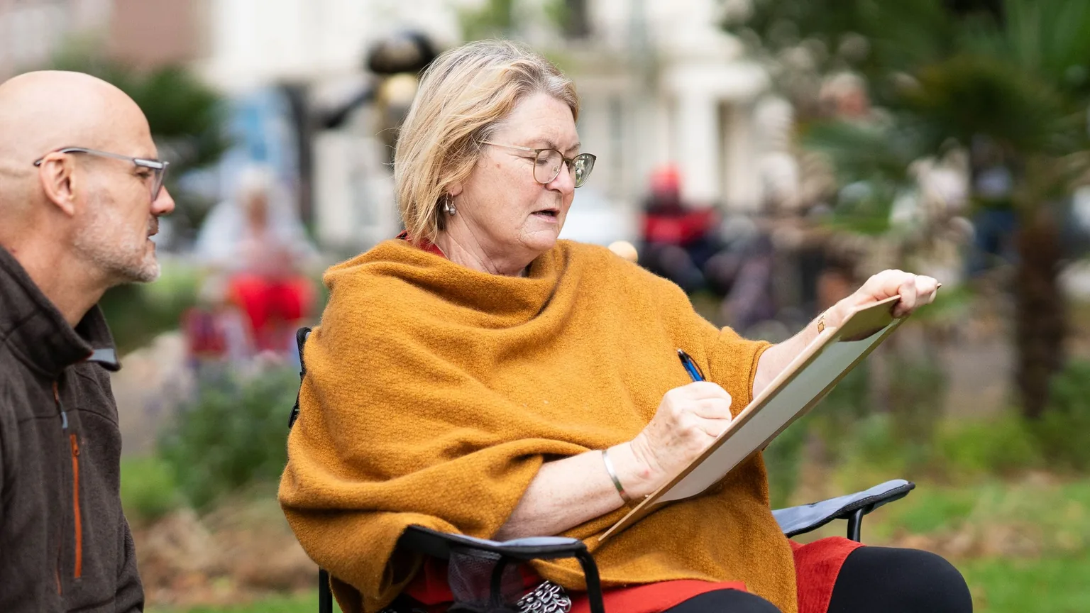 An older woman sketching in a park, with another sketcher beside her