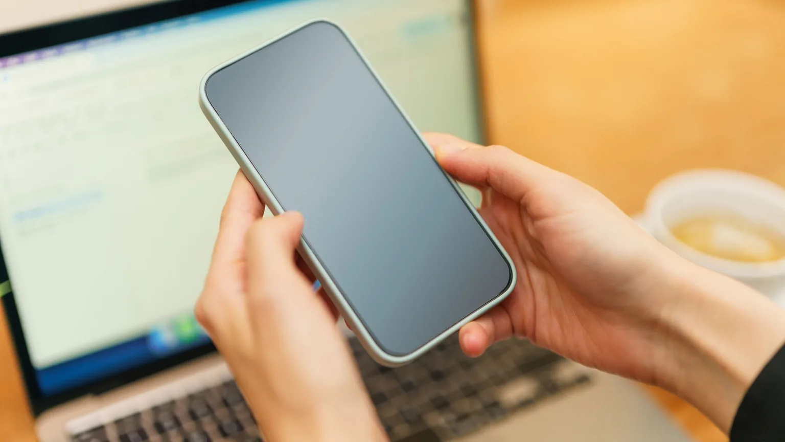 A hand holding a phone above a laptop at a co-working desk