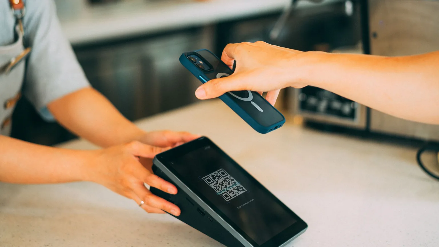 A member tapping a phone on a payment terminal showing a QR code