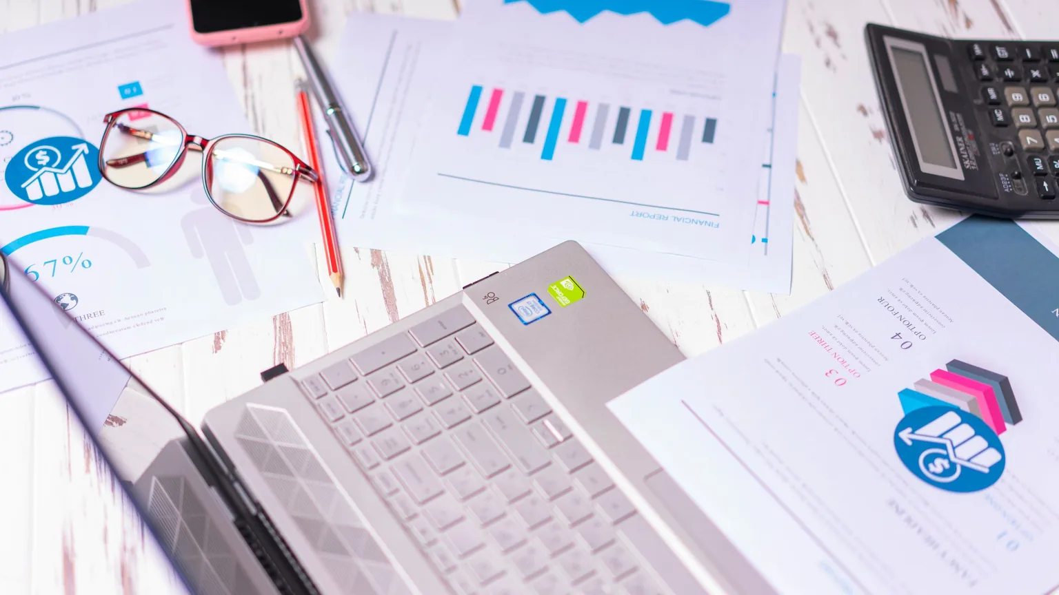 A desk covered in printed reports, a laptop, a calculator and a phone