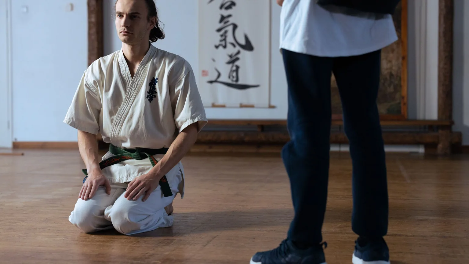 A martial arts student kneeling in the dojo as a visitor arrives